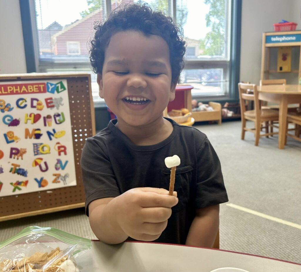 Photo of preschool kid smiling at snack time holding pretzel and marshmallow